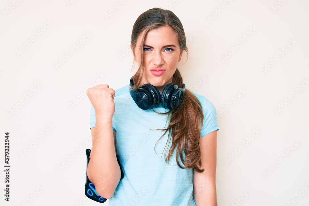 Fototapeta premium Beautiful young caucasian woman wearing gym clothes and using headphones angry and mad raising fist frustrated and furious while shouting with anger. rage and aggressive concept.