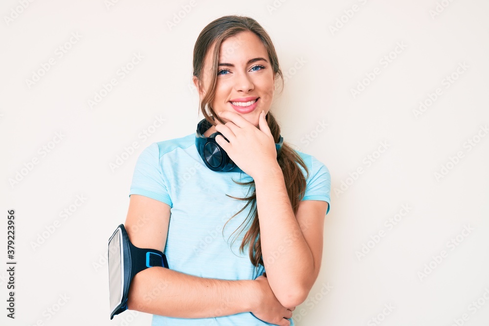 Beautiful young caucasian woman wearing gym clothes and using headphones looking confident at the camera smiling with crossed arms and hand raised on chin. thinking positive. Beautiful young caucasian woman wearing gym clothes and using headphones looking confident at the camera smiling with crossed arms and hand raised on chin. thinking positive.