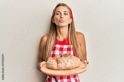 Young blonde woman wearing baker uniform holding homemade bread looking at the camera blowing a kiss being lovely and sexy. love expression.