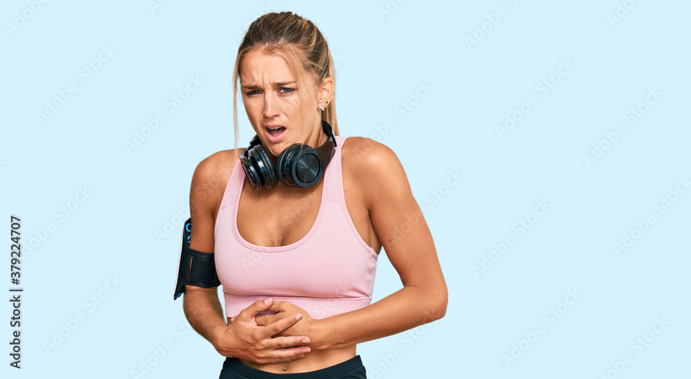 Naklejka premium Young blonde woman wearing gym clothes and using headphones with hand on stomach because nausea, painful disease feeling unwell. ache concept.