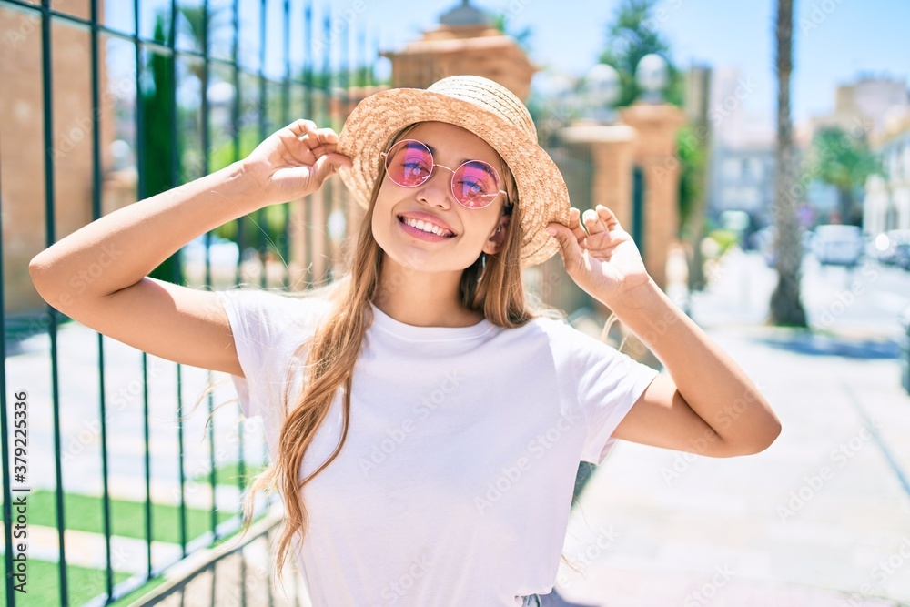 Young beautiful blonde caucasian woman smiling happy outdoors on a sunny day wearing summer hat and pink sunglasses