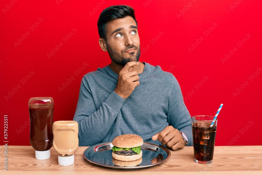 Handsome hispanic man eating a tasty classic burger and soda thinking concentrated about doubt with finger on chin and looking up wondering Handsome hispanic man eating a tasty classic burger and soda thinking concentrated about doubt with finger on chin and looking up wondering