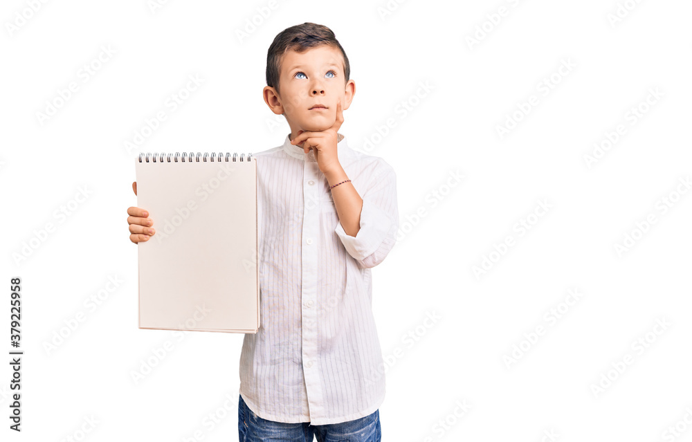 Cute blond kid holding notebook serious face thinking about question with hand on chin, thoughtful about confusing idea Cute blond kid holding notebook serious face thinking about question with hand on chin, thoughtful about confusing idea