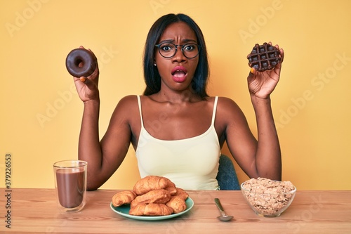 Young african american woman holding chocolate donut and waffle for breakfast in shock face, looking skeptical and sarcastic, surprised with open mouth photo