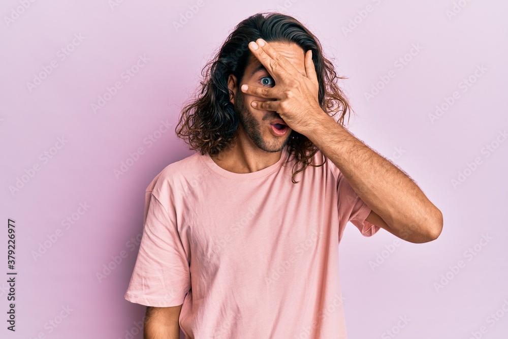 Young handsome man with long hair wearing casual clothes peeking in shock covering face and eyes with hand, looking through fingers afraid