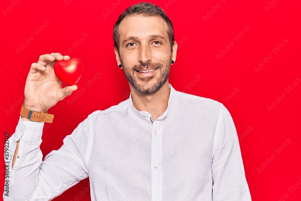 Young handsome man holding heart looking positive and happy standing and smiling with a confident smile showing teeth Young handsome man holding heart looking positive and happy standing and smiling with a confident smile showing teeth