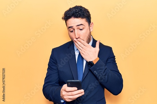 Young hispanic man wearing suit using smartphone covering mouth with hand, shocked and afraid for mistake. surprised expression © Krakenimages.com
