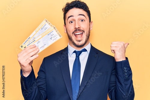 Fotografie Young hispanic man wearing suit holding boarding pass pointing thumb up to the s