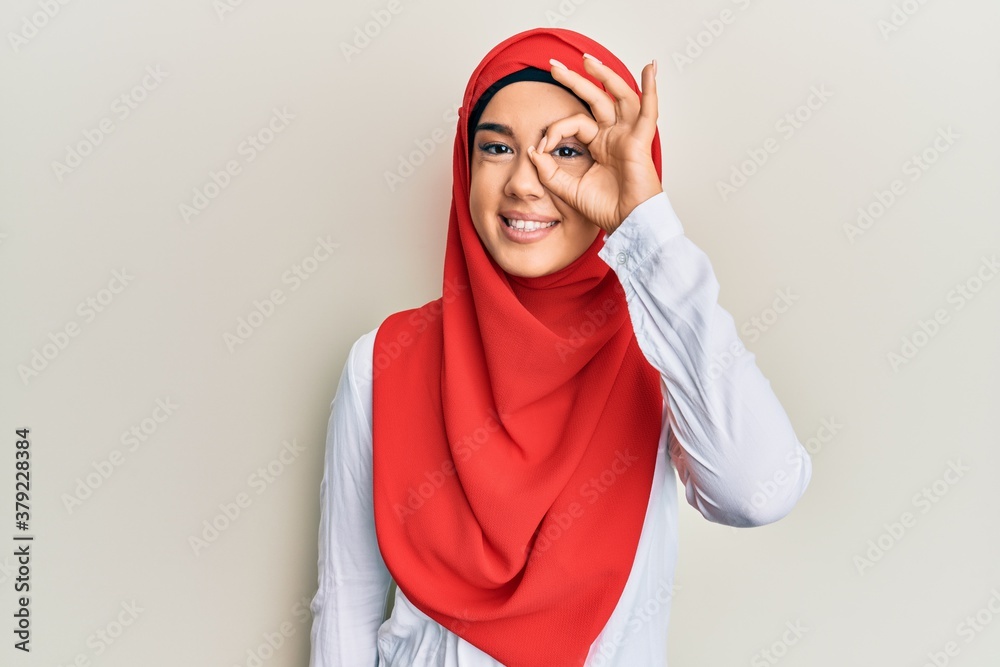 Young beautiful hispanic girl wearing traditional islamic hijab scarf doing ok gesture with hand smiling, eye looking through fingers with happy face. Young beautiful hispanic girl wearing traditional islamic hijab scarf doing ok gesture with hand smiling, eye looking through fingers with happy face.