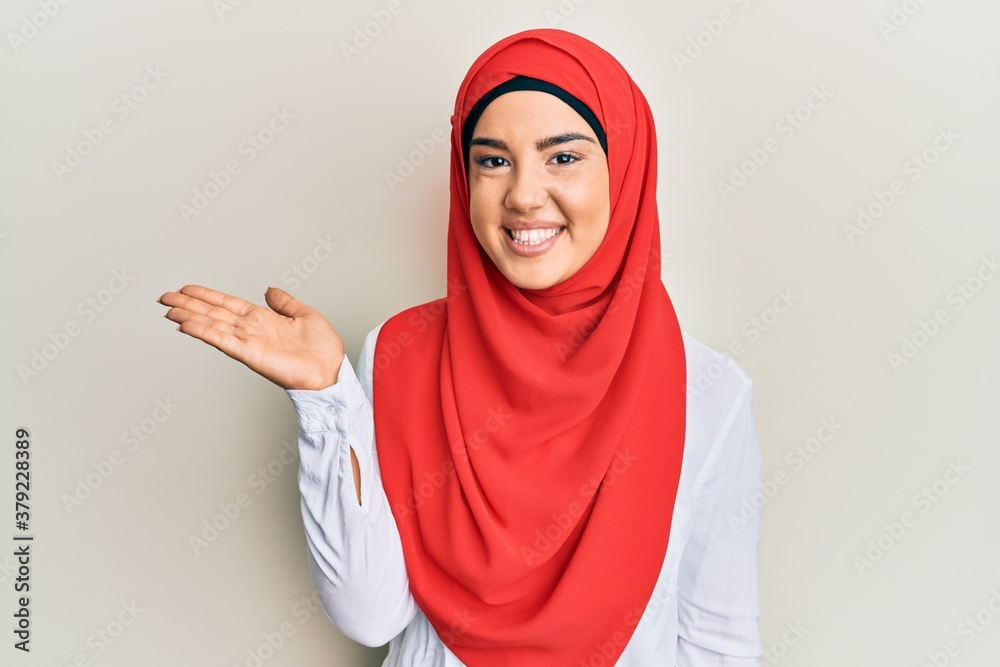 Young beautiful hispanic girl wearing traditional islamic hijab scarf smiling cheerful presenting and pointing with palm of hand looking at the camera. Young beautiful hispanic girl wearing traditional islamic hijab scarf smiling cheerful presenting and pointing with palm of hand looking at the camera.