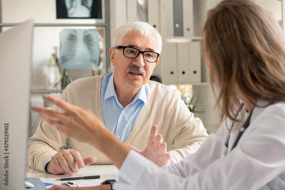 Senior patient in eyeglasses looking at doctor pointing at computer screen Senior patient in eyeglasses looking at doctor pointing at computer screen