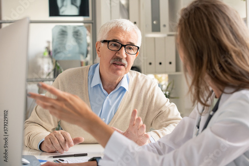 Senior patient in eyeglasses looking at doctor pointing at computer screen