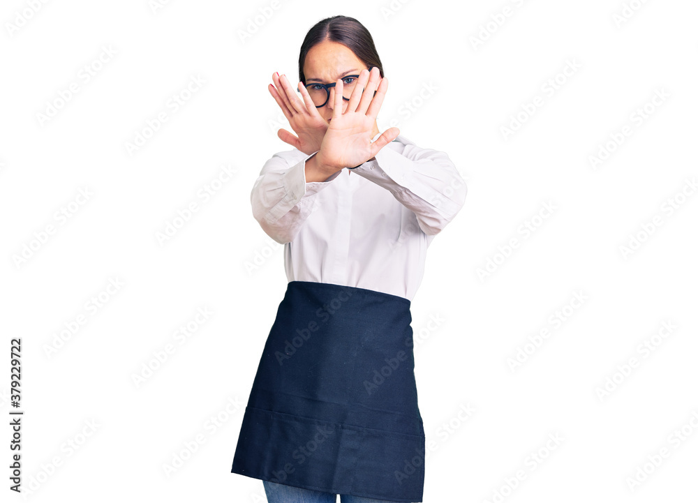 Beautiful brunette young woman wearing professional waitress apron rejection expression crossing arms doing negative sign, angry face Beautiful brunette young woman wearing professional waitress apron rejection expression crossing arms doing negative sign, angry face