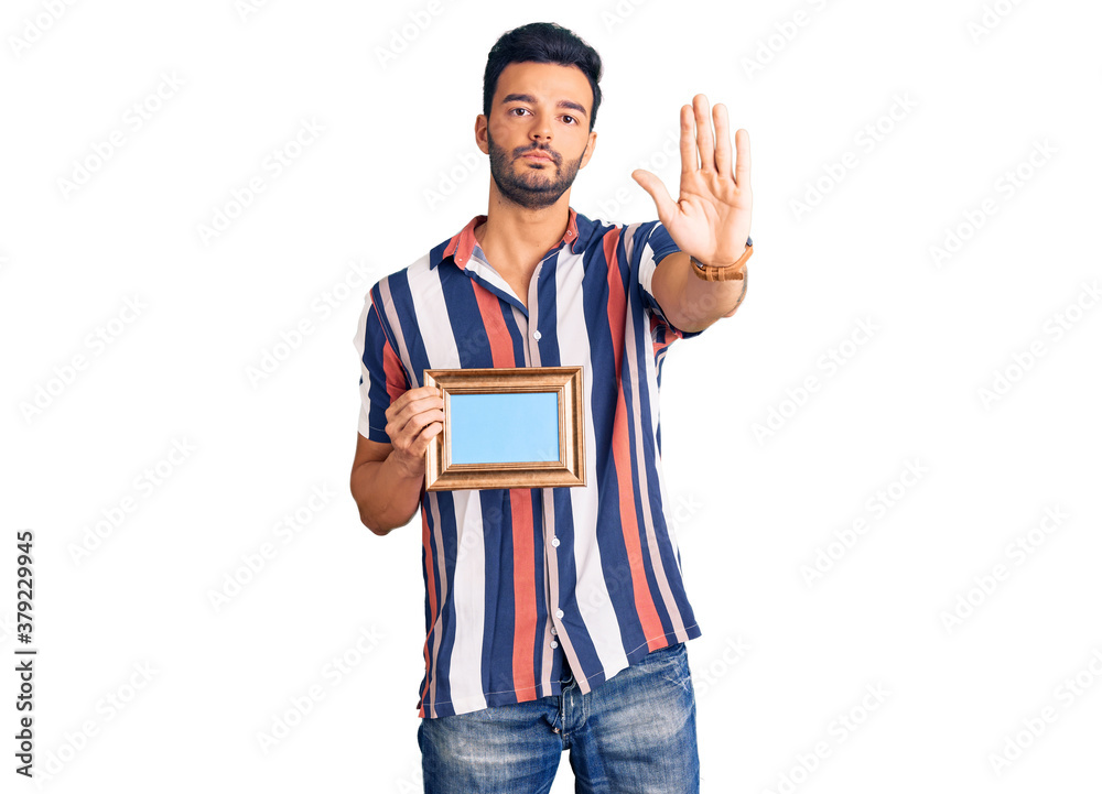 Young handsome hispanic man holding empty frame with open hand doing stop sign with serious and confident expression, defense gesture Young handsome hispanic man holding empty frame with open hand doing stop sign with serious and confident expression, defense gesture