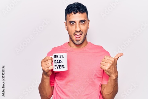 Young hispanic man drinking mug of coffe with best dad ever message pointing thumb up to the side smiling happy with open mouth