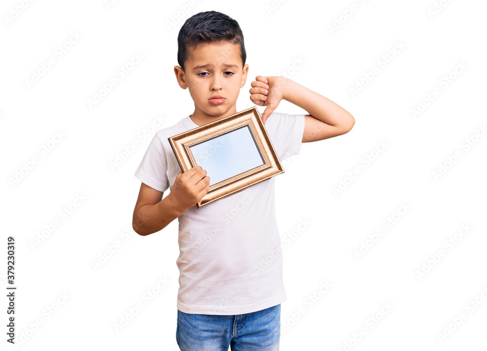 Little cute boy kid holding empty frame with angry face, negative sign showing dislike with thumbs down, rejection concept Little cute boy kid holding empty frame with angry face, negative sign showing dislike with thumbs down, rejection concept