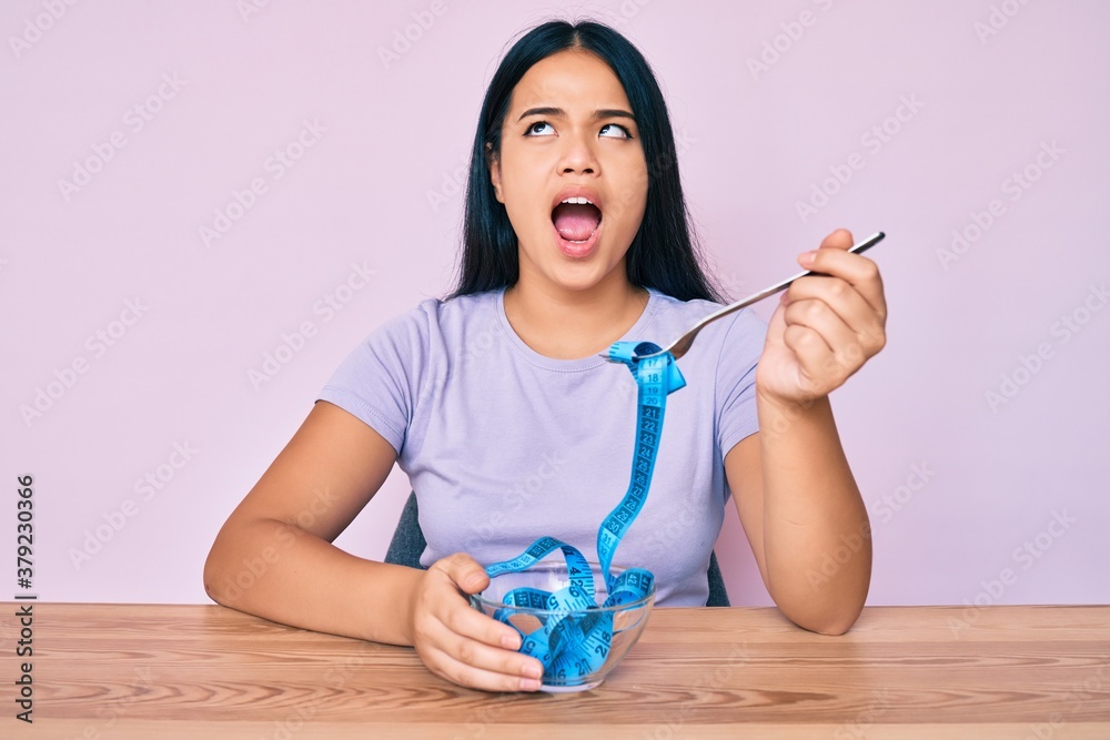 Φωτογραφία Young asian girl eating measure meter doing diet for weight loss angry and mad screaming frustrated and furious, shouting with anger looking up