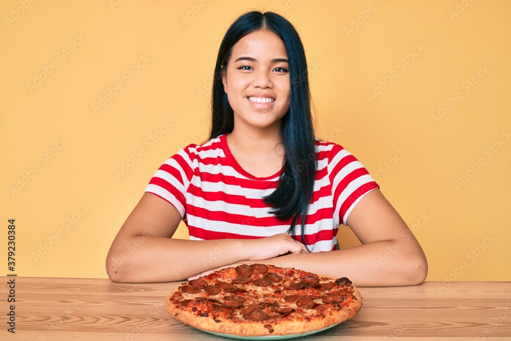 Young beautiful asian girl eating tasty pepperoni pizza looking positive and happy standing and smiling with a confident smile showing teeth Young beautiful asian girl eating tasty pepperoni pizza looking positive and happy standing and smiling with a confident smile showing teeth