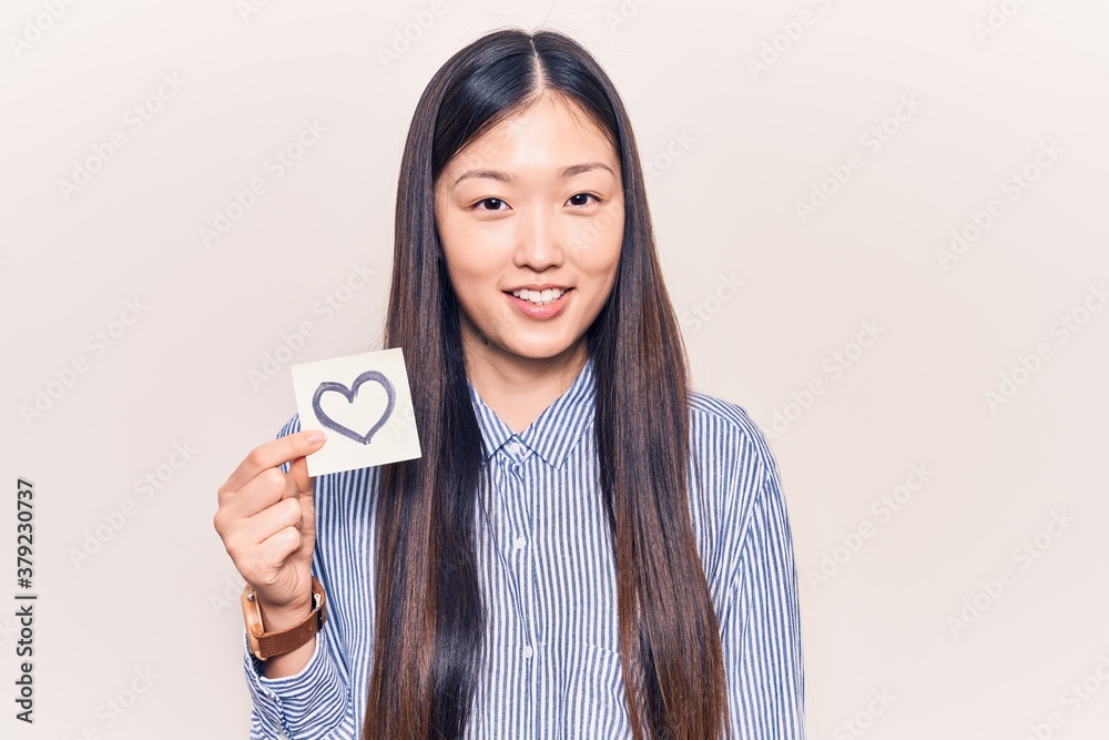 Young beautiful chinese woman holding reminder with heart shape looking positive and happy standing and smiling with a confident smile showing teeth Young beautiful chinese woman holding reminder with heart shape looking positive and happy standing and smiling with a confident smile showing teeth