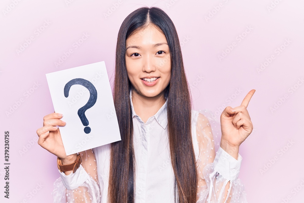 Young beautiful chinese woman holding question mark smiling happy pointing with hand and finger to the side Young beautiful chinese woman holding question mark smiling happy pointing with hand and finger to the side