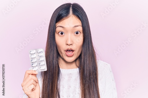 Foto Young beautiful chinese woman holding pills scared and amazed with open mouth fo