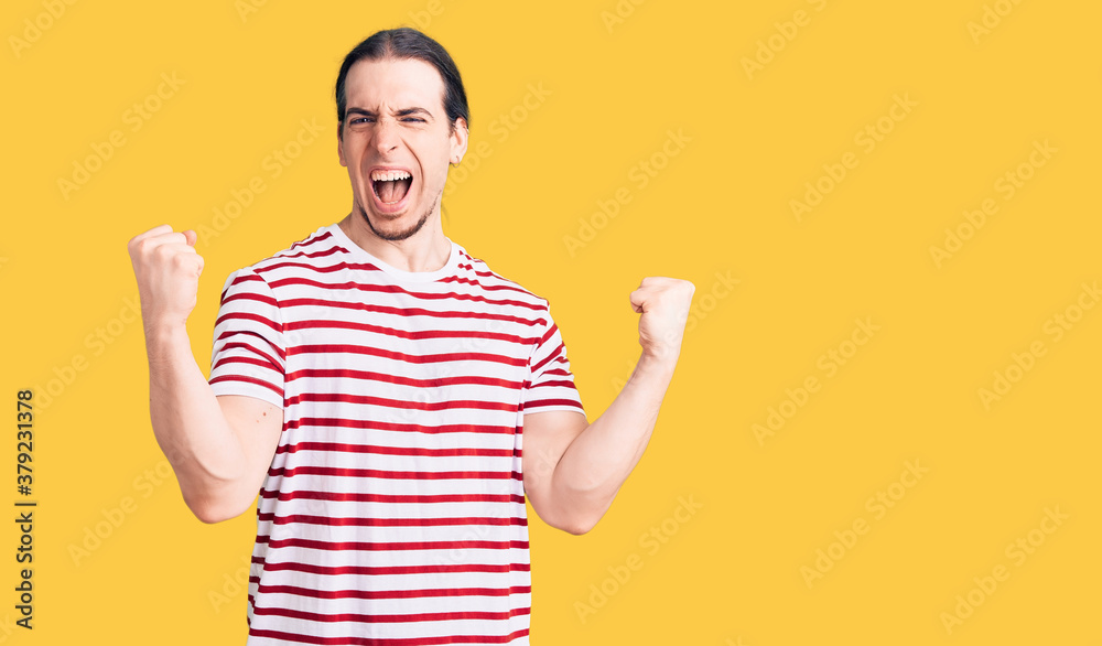 Young adult man with long hair wearing casual striped tshirt screaming proud, celebrating victory and success very excited with raised arms