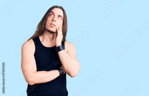 Young adult man with long hair wearing rocker style with black clothes and contact lenses serious face thinking about question with hand on chin, thoughtful about confusing idea © Krakenimages.com