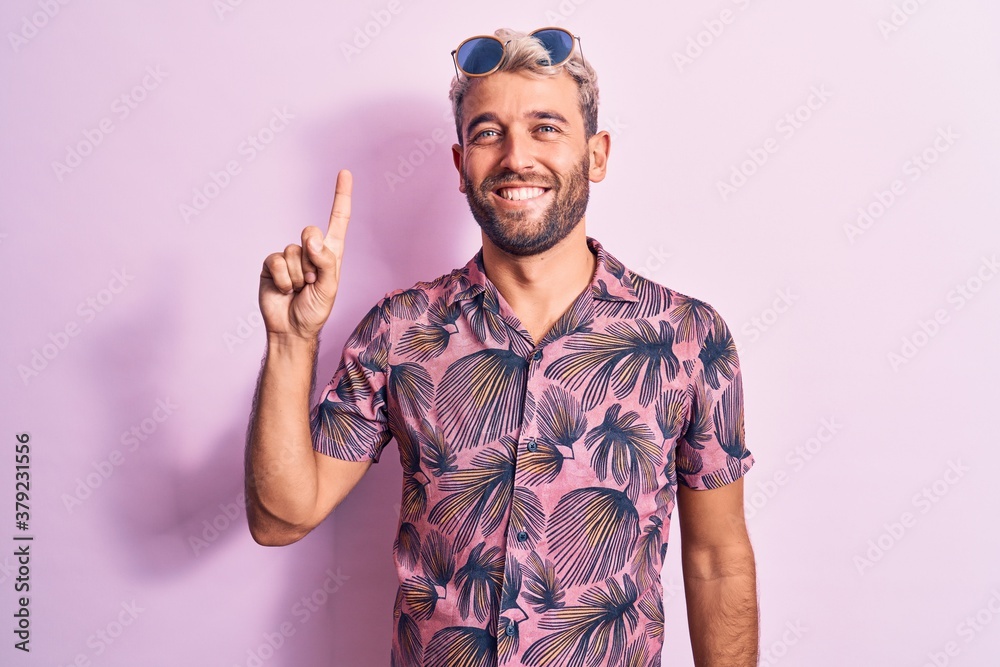Handsome blond man on vacation wearing casual shirt and sunglasses over pink background showing and pointing up with finger number one while smiling confident and happy. Handsome blond man on vacation wearing casual shirt and sunglasses over pink background showing and pointing up with finger number one while smiling confident and happy.