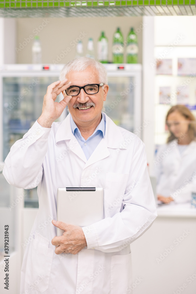 Successful senior drugstore worker in whitecoat and eyeglasses using tablet Successful senior drugstore worker in whitecoat and eyeglasses using tablet