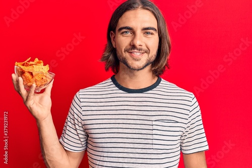 Young handsome man holding nachos potato chips looking positive and happy standing and smiling with a confident smile showing teeth