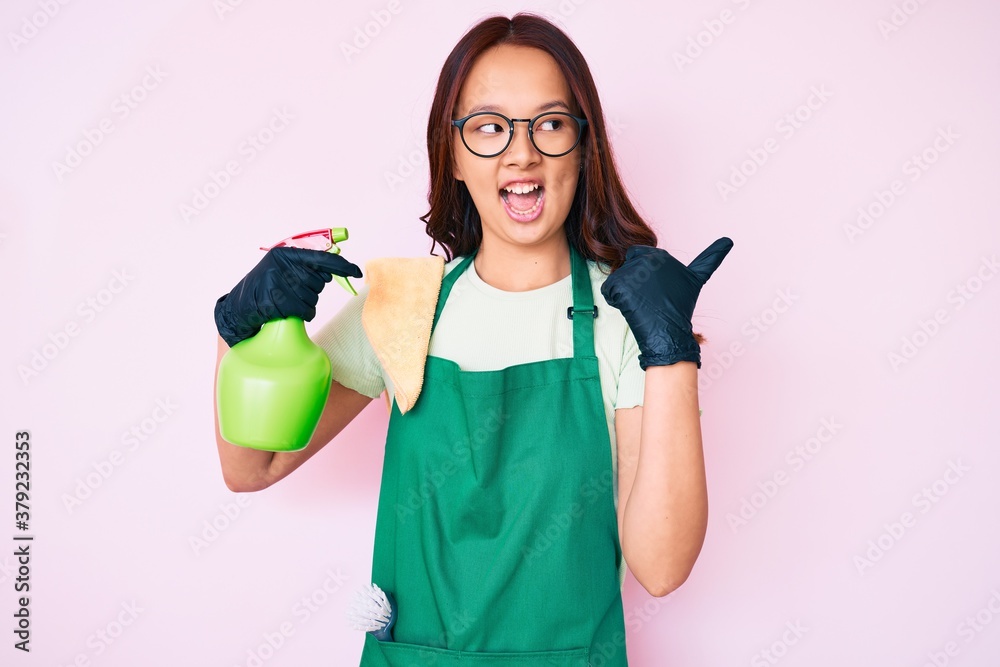 Young beautiful chinese girl wearing apron holding sprayer pointing thumb up to the side smiling happy with open mouth Young beautiful chinese girl wearing apron holding sprayer pointing thumb up to the side smiling happy with open mouth