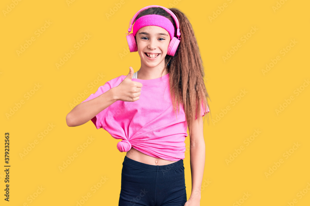 Cute hispanic child girl wearing gym clothes and using headphones doing happy thumbs up gesture with hand. approving expression looking at the camera showing success. Cute hispanic child girl wearing gym clothes and using headphones doing happy thumbs up gesture with hand. approving expression looking at the camera showing success.