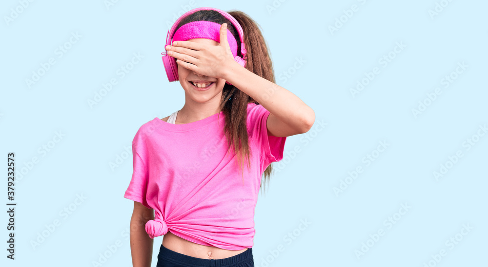 Cute hispanic child girl wearing gym clothes and using headphones smiling and laughing with hand on face covering eyes for surprise. blind concept. Cute hispanic child girl wearing gym clothes and using headphones smiling and laughing with hand on face covering eyes for surprise. blind concept.