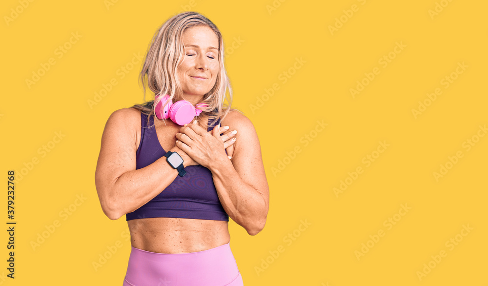 Naklejka premium Middle age fit blonde woman wearing gym clothes and using headphones smiling with hands on chest with closed eyes and grateful gesture on face. health concept.