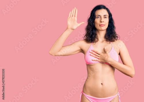 Young beautiful hispanic woman wearing bikini swearing with hand on chest and open palm, making a loyalty promise oath