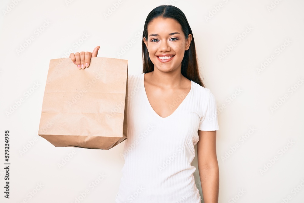 Young beautiful latin girl holding take away paper bag looking positive and happy standing and smiling with a confident smile showing teeth Young beautiful latin girl holding take away paper bag looking positive and happy standing and smiling with a confident smile showing teeth