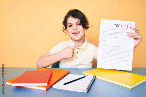 Cute hispanic child showing a passed exam sitting on the desk smiling happy pointing with hand and finger