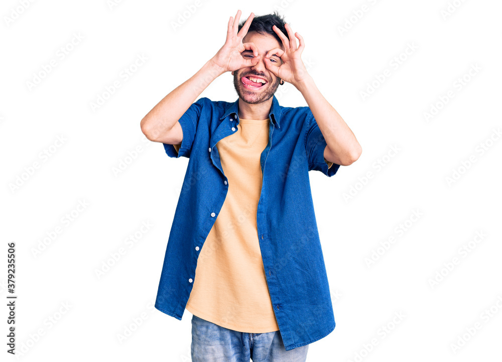 Young hispanic man wearing casual clothes doing ok gesture like binoculars sticking tongue out, eyes looking through fingers. crazy expression. Young hispanic man wearing casual clothes doing ok gesture like binoculars sticking tongue out, eyes looking through fingers. crazy expression.
