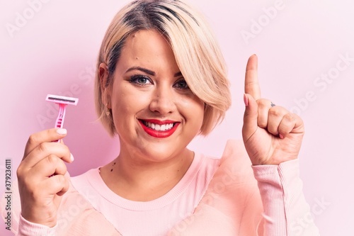 Young beautiful blonde plus size woman holding depilation razor over isolated pink background smiling with an idea or question pointing finger with happy face, number one