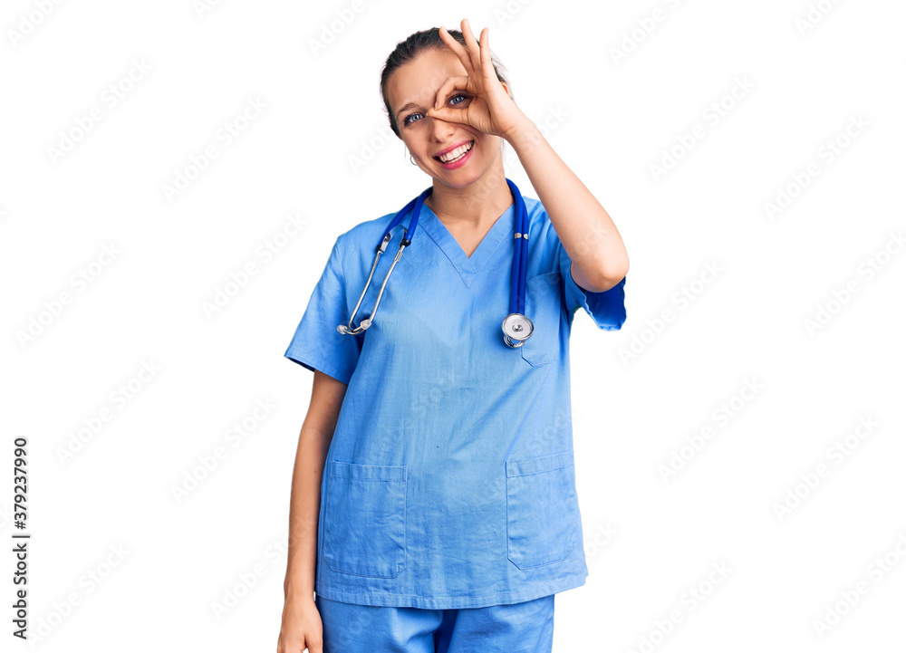 Young beautiful blonde woman wearing doctor uniform and stethoscope doing ok gesture with hand smiling, eye looking through fingers with happy face. Young beautiful blonde woman wearing doctor uniform and stethoscope doing ok gesture with hand smiling, eye looking through fingers with happy face.