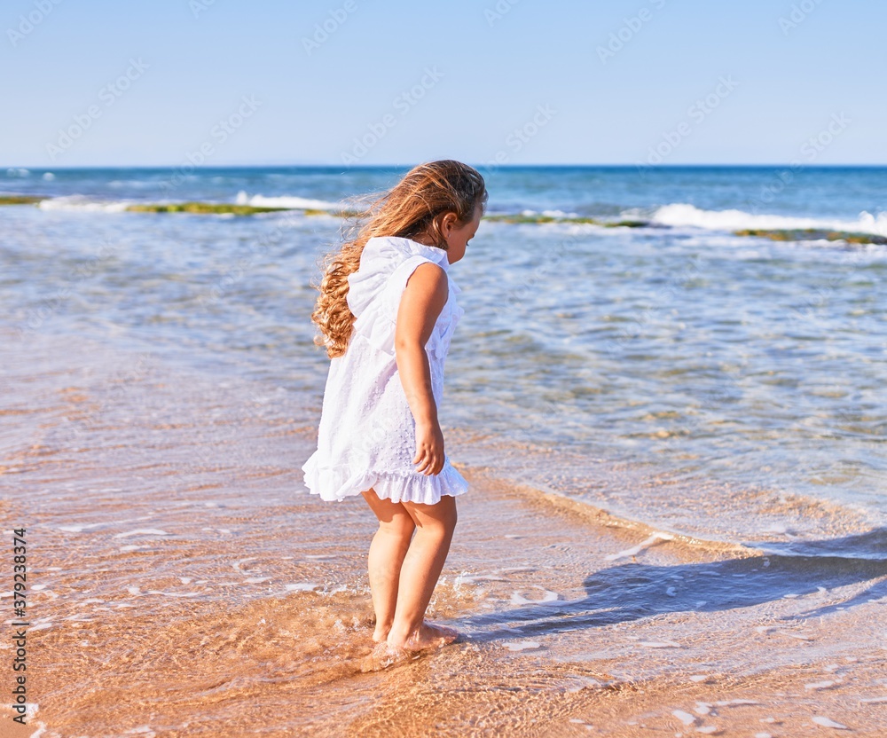 Adorable blonde child on back view wearing summer dress playing on the sand at the beach Adorable blonde child on back view wearing summer dress playing on the sand at the beach