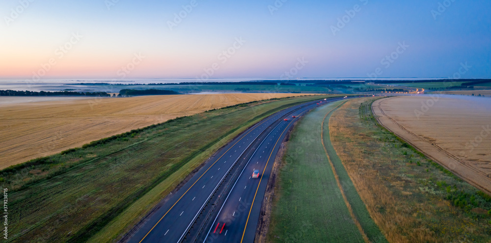 Highway going through meadow in mist surrounded by forest. Aerial view of a road covered in fog.