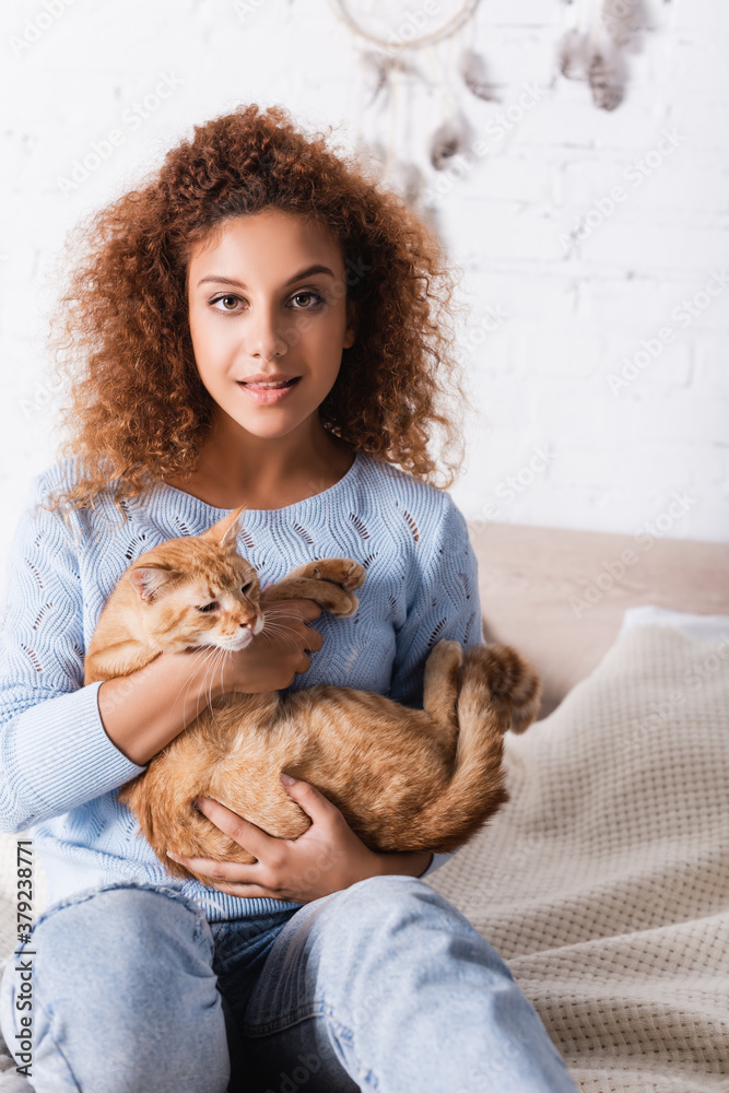 Selective focus of young woman looking at camera while holding tabby cat on bed Selective focus of young woman looking at camera while holding tabby cat on bed