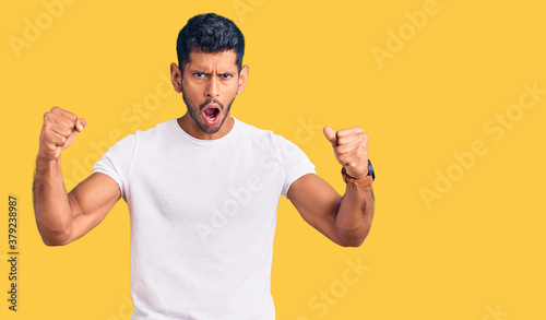 Young latin man wearing casual clothes angry and mad raising fists frustrated and furious while shouting with anger. rage and aggressive concept.
