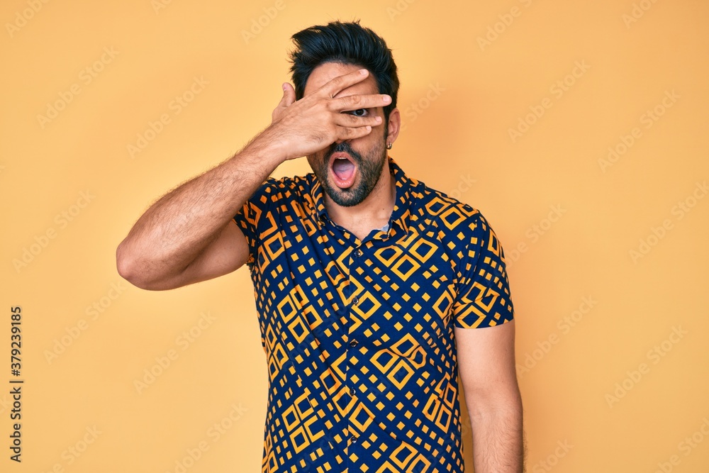 Handsome hispanic man with beard wearing 80s disco style shirt peeking in shock covering face and eyes with hand, looking through fingers with embarrassed expression. Handsome hispanic man with beard wearing 80s disco style shirt peeking in shock covering face and eyes with hand, looking through fingers with embarrassed expression.