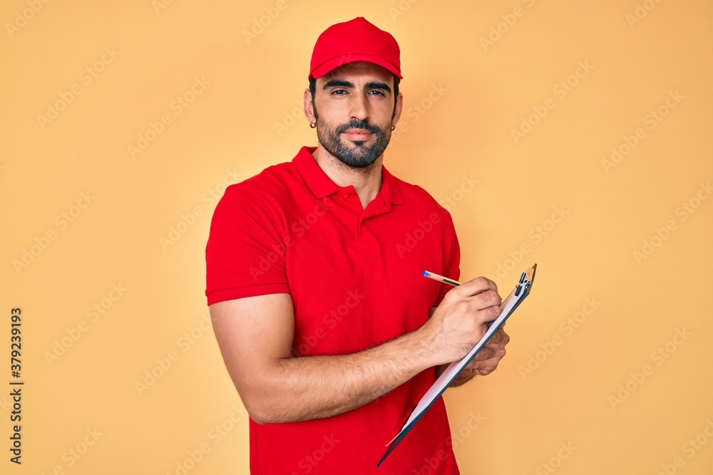 Handsome hispanic delivery man with beard checking list of deliveries relaxed with serious expression on face. simple and natural looking at the camera. Handsome hispanic delivery man with beard checking list of deliveries relaxed with serious expression on face. simple and natural looking at the camera.