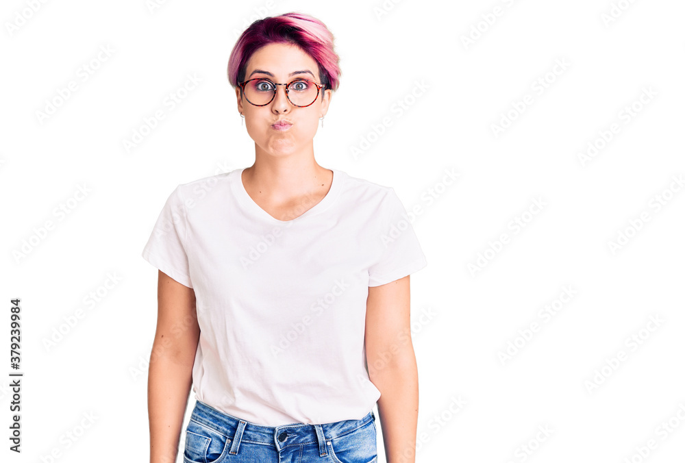 Young beautiful woman with pink hair wearing casual clothes and glasses puffing cheeks with funny face. mouth inflated with air, crazy expression. Young beautiful woman with pink hair wearing casual clothes and glasses puffing cheeks with funny face. mouth inflated with air, crazy expression.