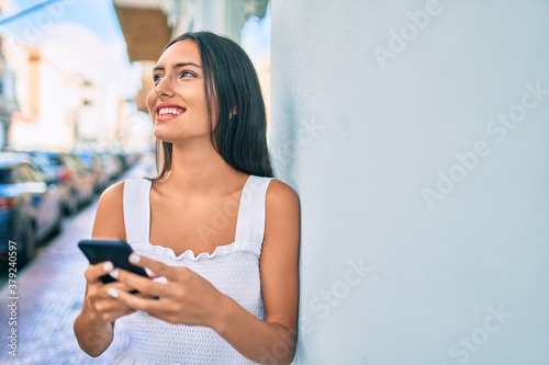 Young latin girl smiling happy using smartphone leaning on the wall. © Krakenimages.com