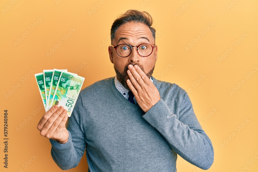 Handsome middle age man holding 50 israel shekels banknotes covering mouth with hand, shocked and afraid for mistake. surprised expression Handsome middle age man holding 50 israel shekels banknotes covering mouth with hand, shocked and afraid for mistake. surprised expression