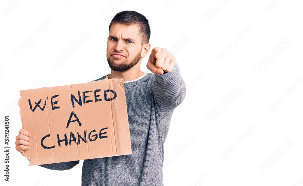 Young handsome man holding we need a change banner pointing with finger to the camera and to you, confident gesture looking serious